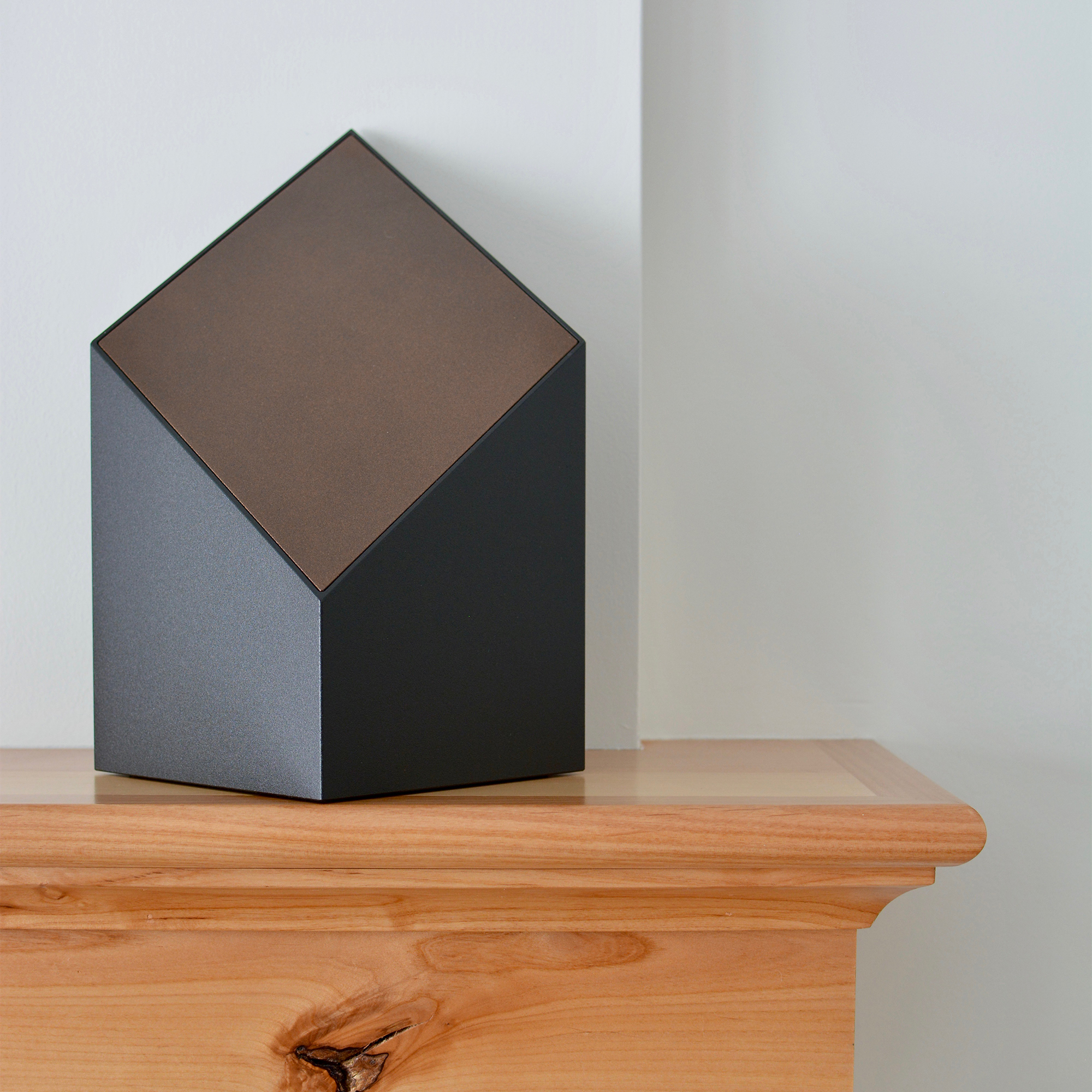 A black everlast series modern cremation urn with a bronze face sitting on a mantel.